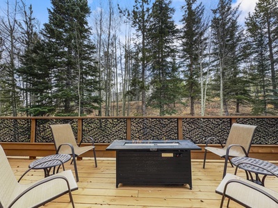 Outdoor balcony with forest view featuring four patio chairs and a fire pit table on a wooden deck. Trees in the background are a mix of evergreen and deciduous.