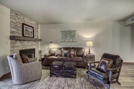 A cozy living room features a brown leather couch, armchairs, a stone fireplace, a world map on the wall, and a lamp on a side table. The area rug and ottomans add texture and warmth to the space.