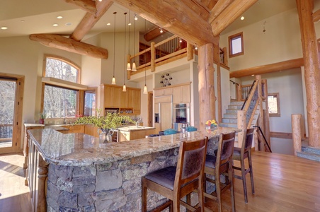 Open-concept kitchen with stone and wood elements, featuring a large island with seating, modern appliances, and a staircase leading to an upper level. Ample natural light from large windows.