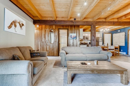 A cozy living room with a wooden ceiling features two beige sofas, a wooden coffee table, and wall art. The dining area and kitchen are visible in the background.