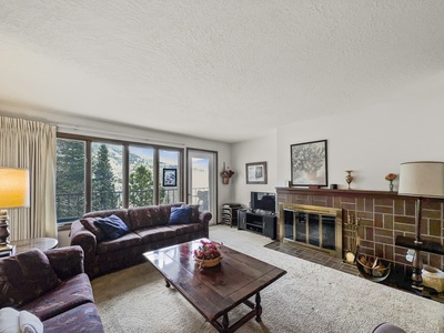 A living room with a large window, two couches, a wooden coffee table, a fireplace, a painting above the mantel, and a television in the corner.