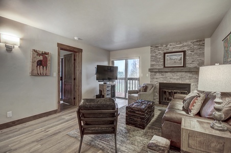 A living room with a TV, chair, sofa, ottoman, and fireplace. The room features a painting of a deer, a stone accent wall, and glass doors leading to an outside view.