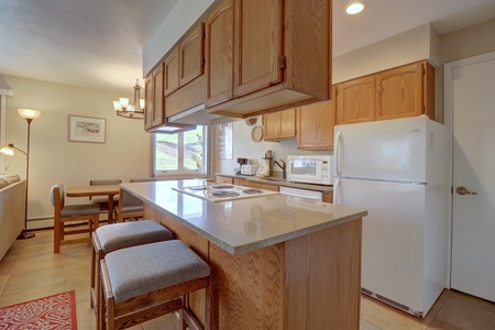 A kitchen with wooden cabinets, a white refrigerator, microwave, and a countertop that extends into a breakfast bar with two stools. A dining table and chairs are in the background near a window.