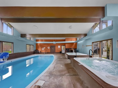 An indoor swimming pool and adjacent hot tub in a well-lit room with large windows, wood beams, and blue and orange walls.