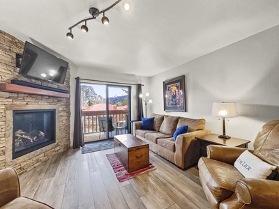 A cozy living room with a fireplace, mounted TV, couch, armchair, and a coffee table. There's a large window with a view of a mountain landscape and a balcony outside.