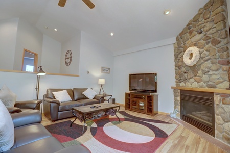 Living room with brown leather sofas, wooden coffee table, flat-screen TV on a stand, and a stone fireplace. The room has a modern decor with a colorful rug and a ceiling fan.