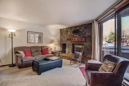 A cozy living room with a brown sofa, chairs, stone fireplace, floor lamp, and large window with a view. The decor includes a framed picture, a throw blanket, and pillows on the seating.