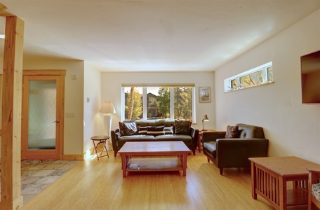 A well-lit living room with wooden flooring, a coffee table, two leather sofas, a side table with a lamp, and large windows offering a view of trees and a neighboring house.