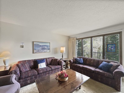 Living room with two purple sofas, wooden coffee table with a flower arrangement, a large window, and a framed picture on the wall. The space is well-lit with natural light.