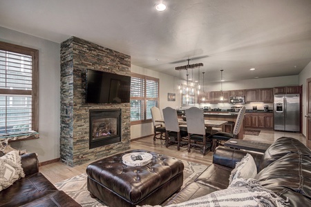 A modern living area with a stone fireplace, wall-mounted TV, and leather seating. The open floor plan extends to a wooden dining set and a kitchen with stainless steel appliances.