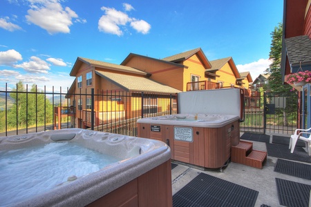 Outdoor hot tubs on a fenced patio with mountain scenery in the background and several multi-story buildings nearby.