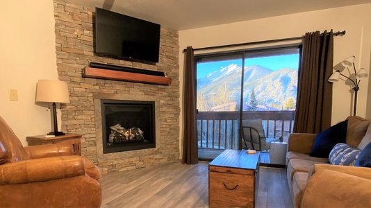 A cozy living room with a stone fireplace and wall-mounted TV, a leather chair, a sofa, and a wooden coffee table. Large window with a mountain view and a balcony. Floor lamp in the corner.