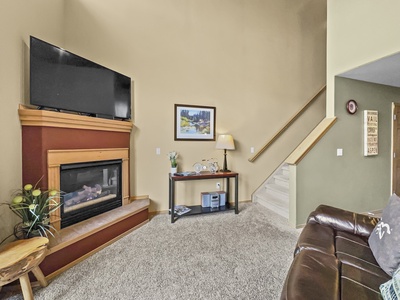 Living room featuring a wall-mounted TV above a fireplace, a small table with decor, a lamp, artwork, and a staircase leading to an upper level. Brown leather couch visible in the foreground.