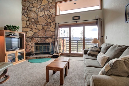 A cozy living room with a stone fireplace, TV stand, two beige sofas, and a wooden coffee table. Large windows with a glass door provide a view of a mountain landscape outside.