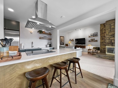 Modern kitchen with bar stools and a countertop overlooking a living room. The living area features a stone fireplace, wall-mounted TV, and assorted furniture, including a lamp and framed pictures.