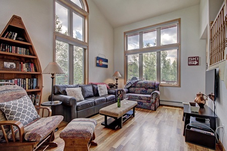 A cozy living room with large windows, two sofas, a wooden bookshelf, a TV, a coffee table, and a decorative "SKI LIFT" sign. The room has wooden flooring and some lamps for lighting.