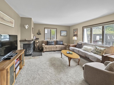 A living room with a flat-screen TV on a wooden stand, two sofas, a fireplace with stacked wood, a coffee table, two large windows, and light-colored carpeting.