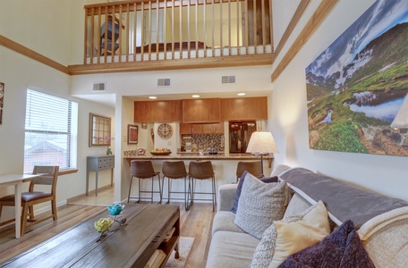 A bright, open-concept living area with a loft and wooden railing. The room features a sofa, coffee table, bar stools at a counter, kitchen, dining area, and wall art. The space has natural light from the windows.