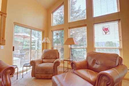 A cozy living room with tall windows, leather armchairs, and a recliner. Outside, there's a balcony with an umbrella table and trees visible through the large glass doors.