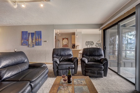 A living room with black leather sofas and a glass coffee table that has a bottle of wine and glasses. There are paintings on the wall, sliding glass doors, and a view of a balcony with outdoor furniture.