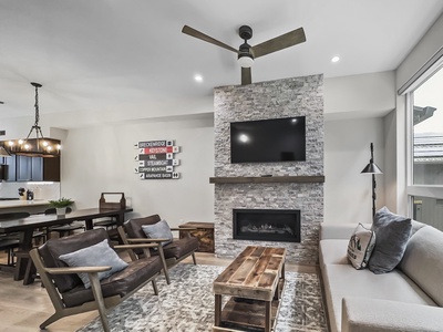 Modern living room featuring a stone fireplace with a mounted TV, ceiling fan, brown chairs, a gray couch, and a wooden coffee table. Dining area and kitchen visible in the background.