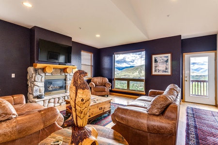 A living room with leather furniture, a stone fireplace, a wooden eagle sculpture, and large windows with mountain views. A wall-mounted TV is above the fireplace and a glass door leads to outside.