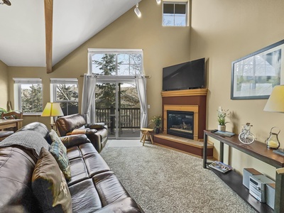 A cozy living room with leather sofas, a fireplace, a wall-mounted TV, and large windows overlooking a balcony with outdoor trees. The room features neutral tones and various decor pieces.