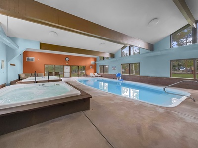 Indoor swimming pool area with blue walls and large windows. The space includes a rectangular swimming pool, two hot tubs, lounge chairs, and a clock on the wall.