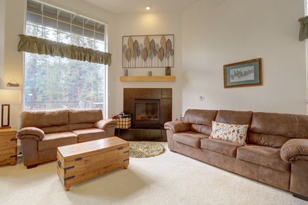 A cozy living room with two brown sofas, a wooden chest as a coffee table, a fireplace with a modern wood art piece above it, and large windows bringing in natural light with views of trees outside.