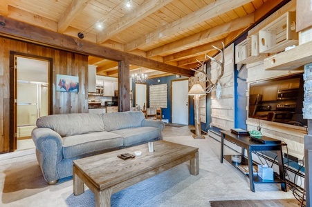 A cozy living room with a gray sofa, wooden coffee table, mounted deer antlers, and an adjoining kitchen. Exposed wooden beams on the ceiling add to the rustic atmosphere.