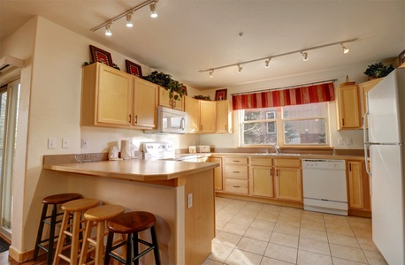 A kitchen with light wood cabinets, a countertop with three wooden stools, white appliances including a refrigerator, stove, and dishwasher, and a window with a red striped valance.