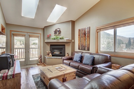 A cozy living room features a leather sofa, armchair, wooden coffee table, and TV. Skylights and a large window illuminate the space, which includes a fireplace and decor of forest-themed art and figures.