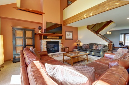 Living room with a large sectional sofa, stone fireplace, wall-mounted TV, and wooden furniture. Stairs lead to an upper level, and a dining area is visible in the background.