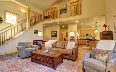 A cozy living room with a high ceiling, featuring two armchairs, a leather sofa, a wooden coffee table, and a staircase leading to an upper floor. The kitchen is visible in the background.