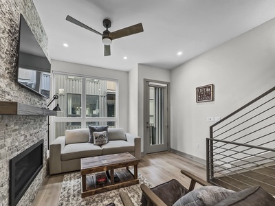 Modern living room with a ceiling fan, beige sofa, wooden coffee table, wall-mounted TV, and a fireplace. A door leads outside, and a staircase is visible on the right. Walls are decorated minimally.