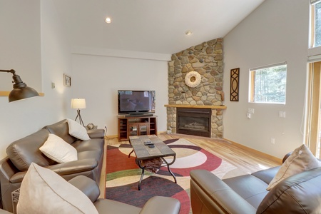 A cozy living room with a stone fireplace, TV on a wooden stand, leather sofas, and a modern rug. Large windows allow natural light in.