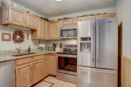 A kitchen with wooden cabinets, a countertop, and stainless steel appliances including a refrigerator, microwave, and oven. There is a small wreath on the wall and decorative items above the cabinets.