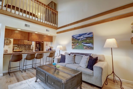 A cozy living room features a beige sofa with cushions, a wooden coffee table, barstools at a kitchen counter, and two floor lamps. A loft with a wooden railing overlooks the space.