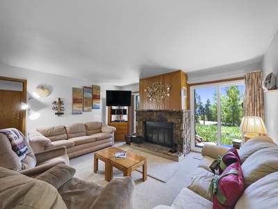 A living room with a stone fireplace, beige furniture, a TV, wooden coffee table, wall art, and sliding glass doors opening to a grassy outdoor area.