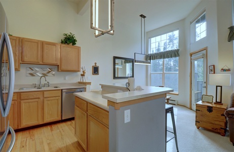 Spacious kitchen with light wood cabinets, stainless steel appliances, and a breakfast bar. Adjacent living area features large windows, high ceilings, and a cozy seating arrangement.