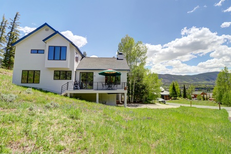 Exterior of home. The home offers a large deck, table with umbrella and chairs, and a grill. The driveway can be seen to the right of the home.