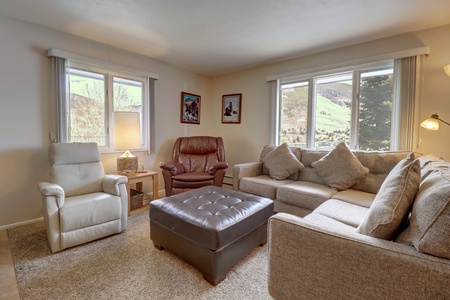 A cozy living room with a beige sectional sofa, two armchairs, a large brown ottoman, and two windows offering views of a scenic landscape. The room is lit by a table lamp and wall-mounted light.