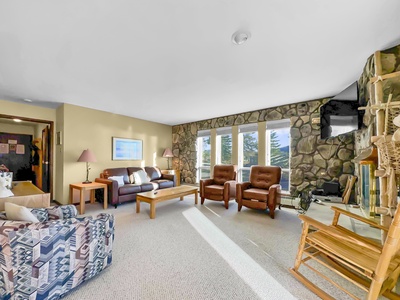 A cozy living room with stone walls, large windows, a sofa, two recliners, a rocking chair, and a wooden coffee table. A TV is mounted above a stone fireplace. Light floods the room from the windows.