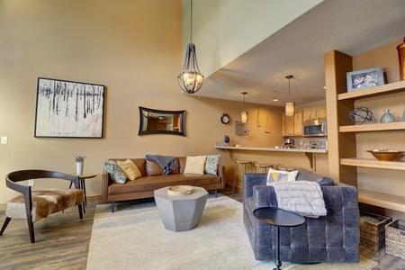 A modern living room with a high ceiling, featuring a brown sofa, a gray armchair, a hexagonal coffee table, and a kitchen in the background. The room is decorated with contemporary art and light fixtures.