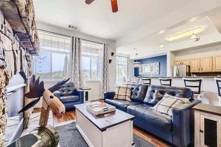 A modern living room with a stone fireplace, blue and white sofas, a coffee table, and large windows. The adjacent kitchen features light wood cabinets, bar seating, and stainless steel appliances.