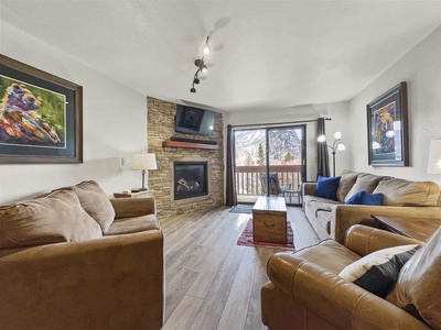 A cozy living room with brown sofas, a fireplace, two paintings of dogs, and a large window with a mountain view. There is also a wooden coffee table, a rug, and a TV mounted above the fireplace.