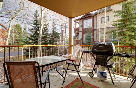 Outdoor patio with a glass dining table, several chairs, and a gas grill. Background shows trees and nearby buildings.