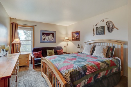 A bedroom with a wooden bed adorned with a colorful patchwork quilt, a dresser on the left, a window with curtains, a sofa with cushions, and mountain-themed wall art.