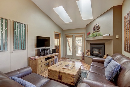 A cozy living room with leather sofas, skylights, a wooden coffee table, wall-mounted TV, and a fireplace. Decor includes paintings, a bookshelf, and potted plants. Door leads to a balcony.