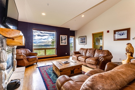 Spacious living room with leather sofas, a large picture window with a mountain view, a stone fireplace, and a wooden coffee table on a red area rug. Decor includes nature-themed art and a carved wooden eagle.